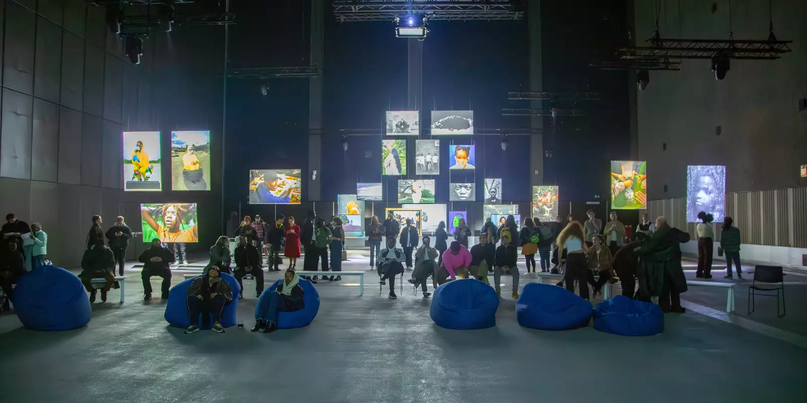 A large group of people in the Reframe exhibition in the Warehouse. They are sat on benches and beanbags looking at the photography.