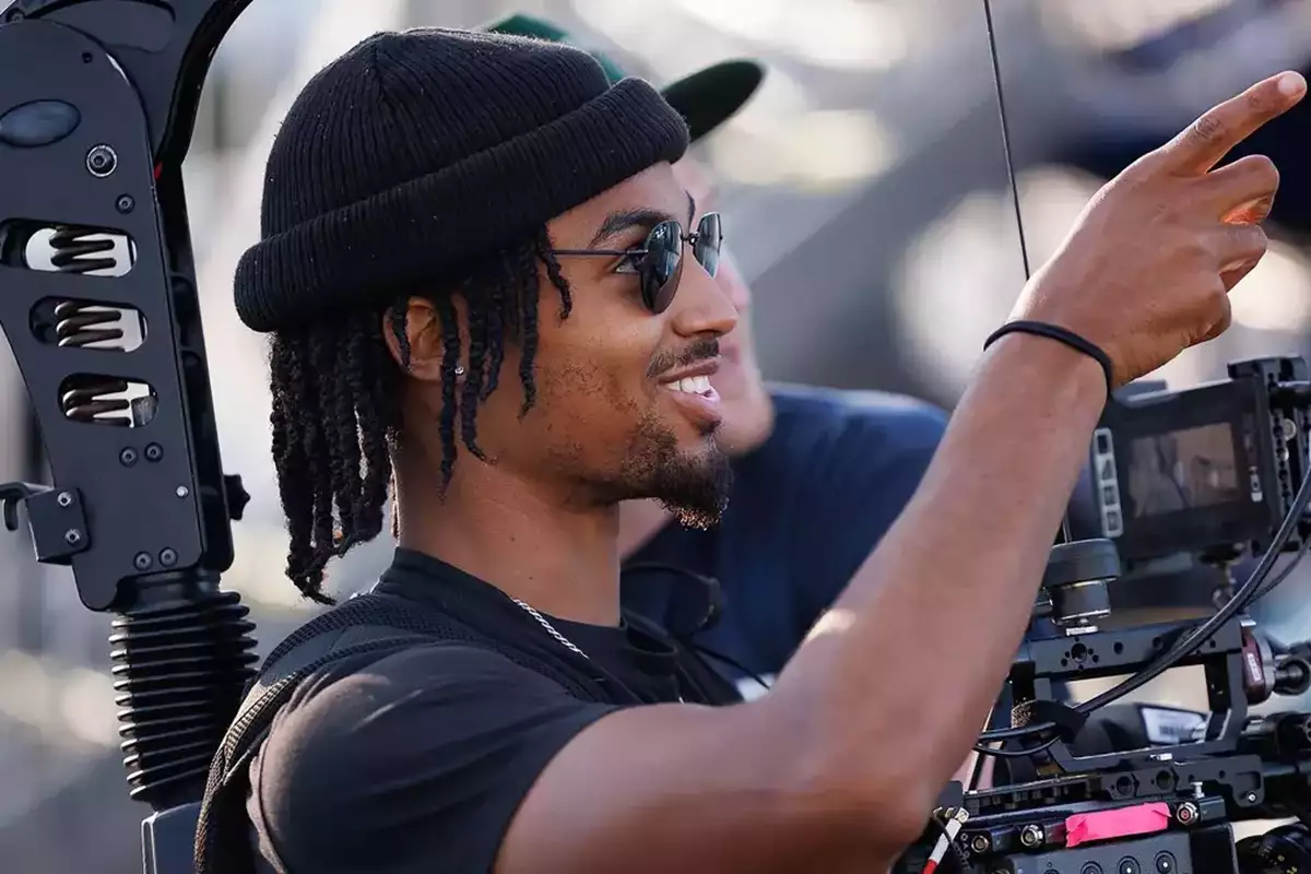 A young Black man wearing sunglasses and a beanie hat operates a video camera.
