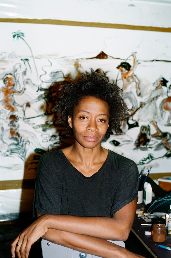 A photographic portrait of a woman artist in a black t-shirt with studio works in the background