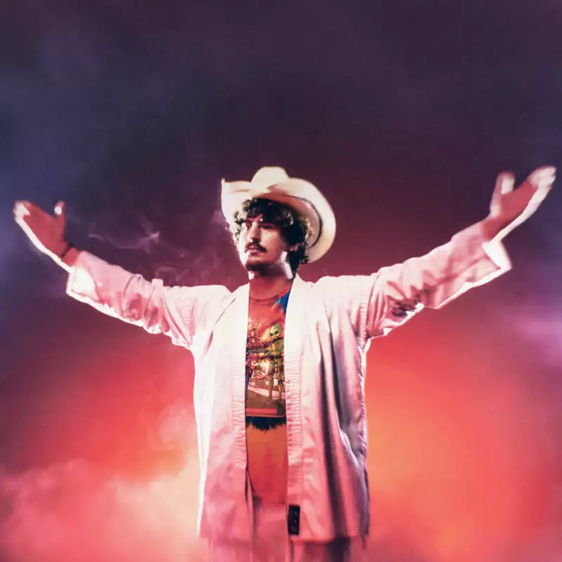 A man in a white suit and cowboy hat stands with arms outstretched, surrounded by dramatic red and blue lighting and thick mist.