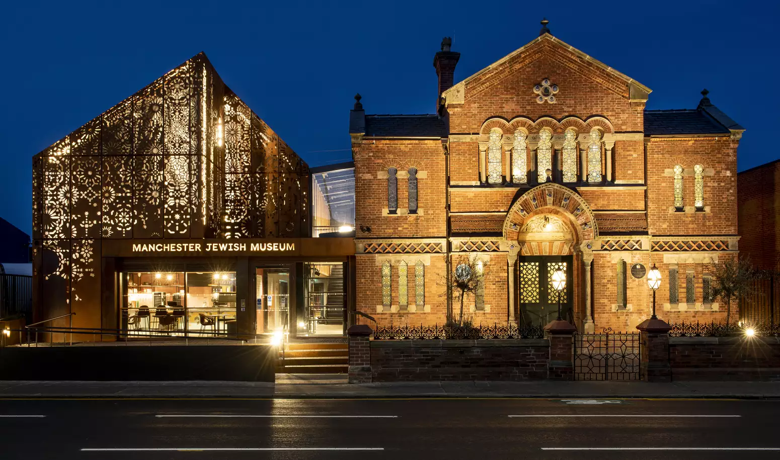 Manchester Jewish Museum seen at night. Lights glow from inside the new extension, which has patterns cut into the metal frontage.
