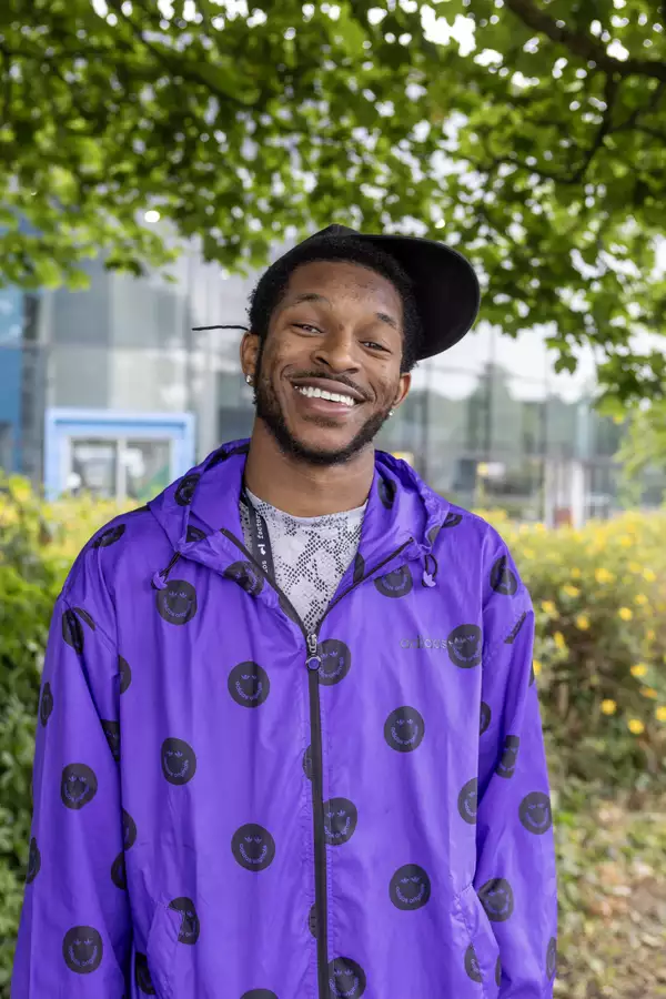 headshot of Zeriah Otutu wearing a black cap and a bright purple waterproof with black smiley faces on it. They are stood outside under a tree.