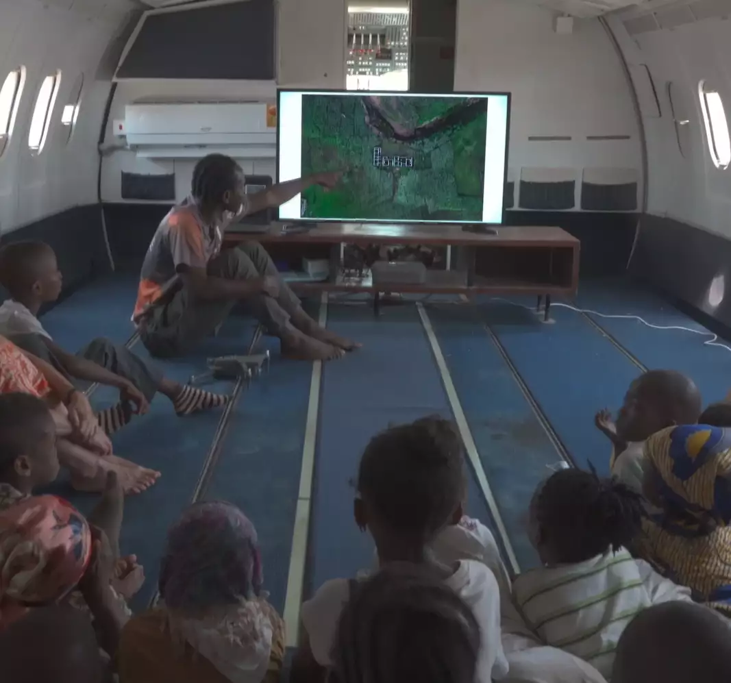 A photograph of a man gesturing at a TV screen showing a map, while delivering a lesson to a group of children in Ghana. The lesson is taking place in a retired airplane with the rows of seats removed.
