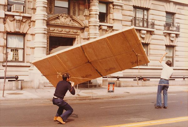 Two men using a pulley system to lower a boxed painting from a tall apartment block