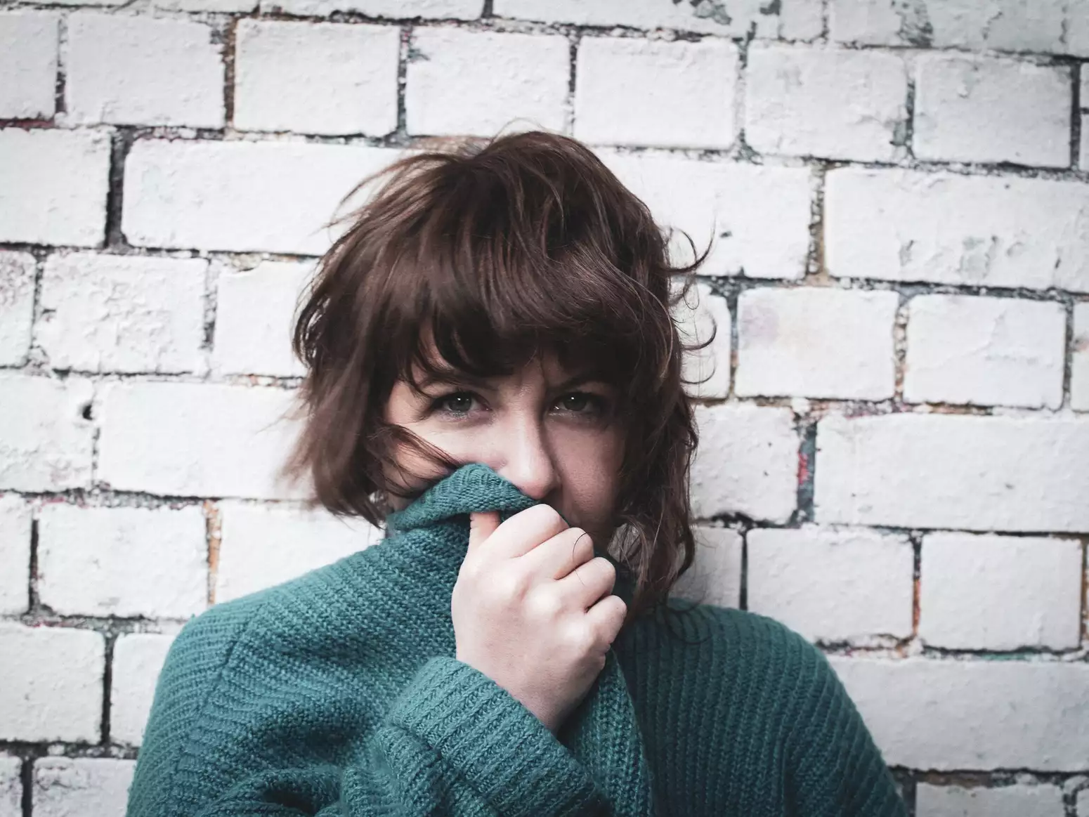 Facing directly into the camera  you can see that Test Card Girl is wearing a green cardigan or jumper, she is partially covering her face with the collar of it. She has wavy brown hair and is standing in front of a white brick wall.