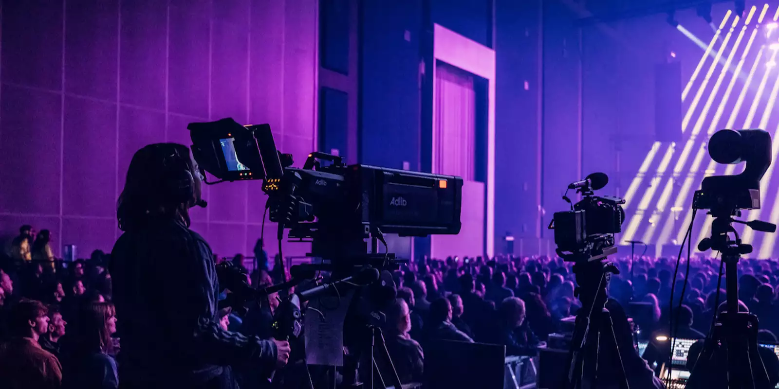 A camera operator films a live event in the Warehouse, which is filled with people. There is dramatic purple lighting with multiple cameras and bright stage beams.