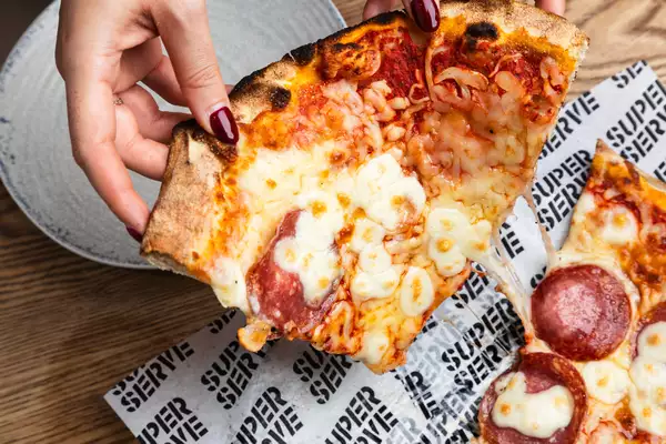 A hand lifting a cheesy slice of pepperoni pizza with a crispy crust over paper that says 'Superserve'.