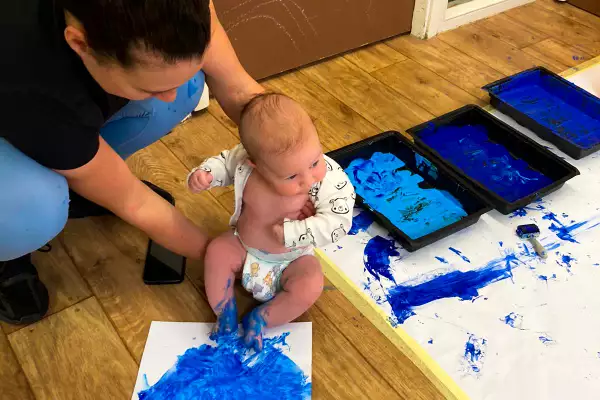 A woman holds a baby with blue paint on its feet. Next to them are sheets of paper with blue paint on them.