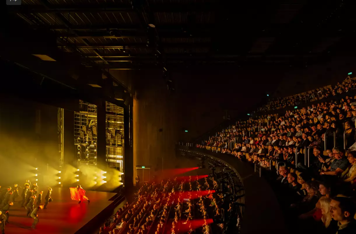 A theatre full of people watching a stage show lit by red and yellow lights