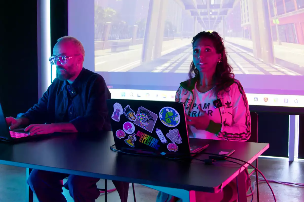 Simon Wilkinson and Myra Appannah sat at a desk on their computers. Behind them is a big screen showing a game.