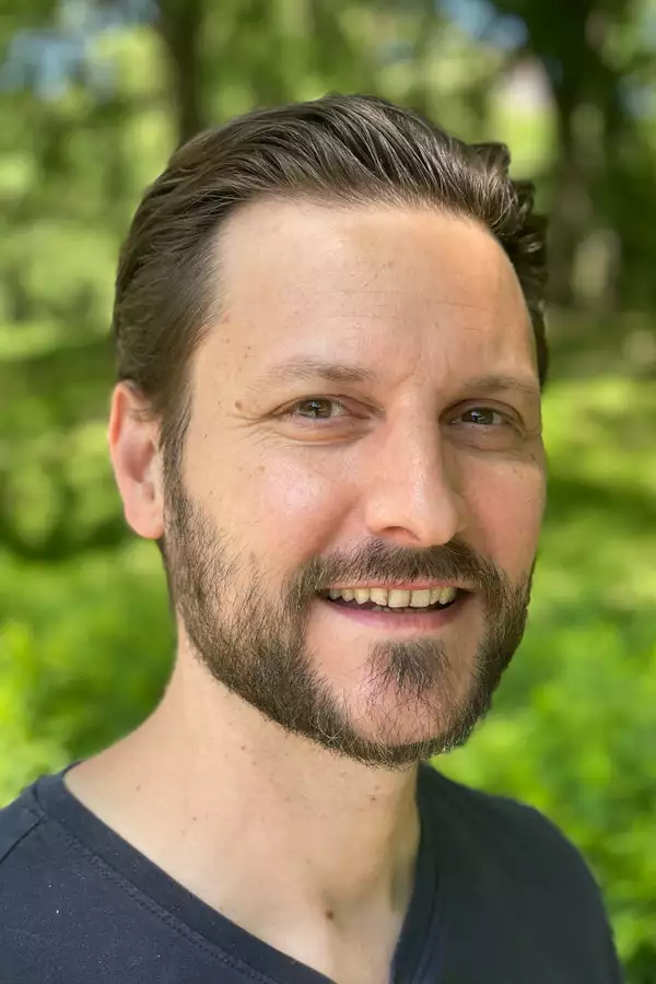 Olly Taylor in a blue t-shirt with greenery behind him