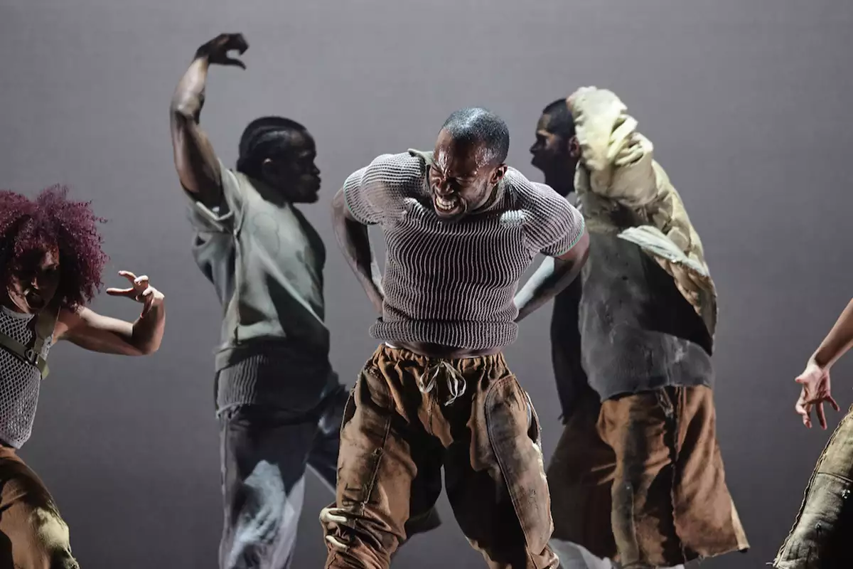 Five contemporary dancers perform with intense, expressive movements on stage, dressed in earthy-toned clothing. The central dancer leads the powerful scene, surrounded by others in dynamic, dramatic poses.