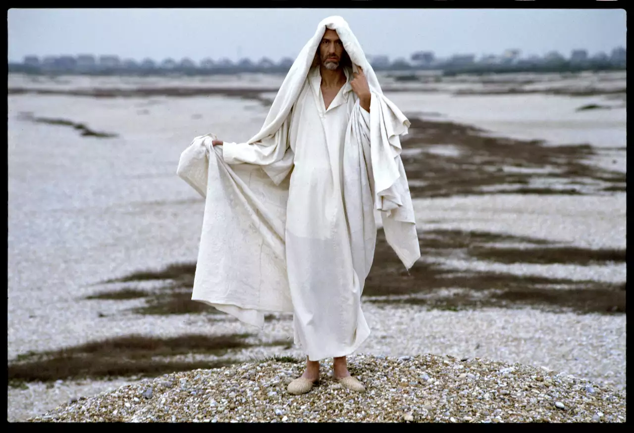 Still from Derek Jarman's The Garden (1990), showing a man dressed in a white robe and headdress stood on a shingle beach.