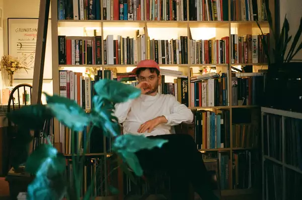 A young man sits in front of a bookcase. He wears a white long-sleeved t-shirt and coral-coloured cap and has dark brown hair. There are plants in the foreground of the image.