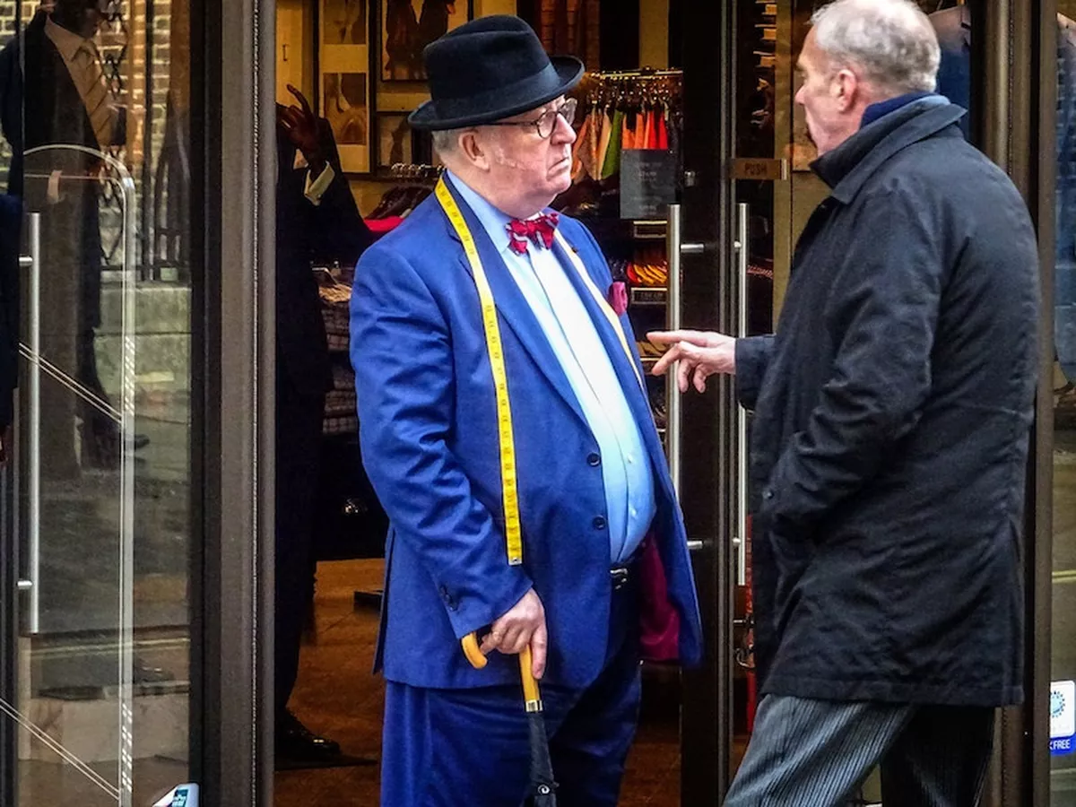 A tailor and a passerby, both of a similar age, deep in conversation outside the tailor's shop