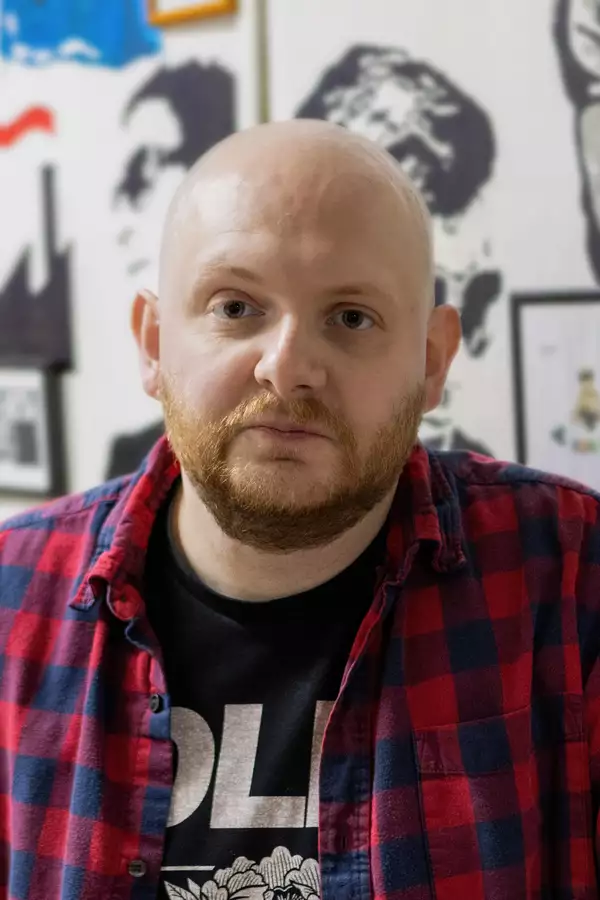 Headshot of Patrick Nicholls wearing a black t-shirt under a red and dark blue chequered open shirt.