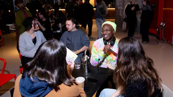A group of people sat around a table in the Social, engaged in conversation and laughter. The table is scattered with drinks, papers, and personal items. In the background are other people chatting.