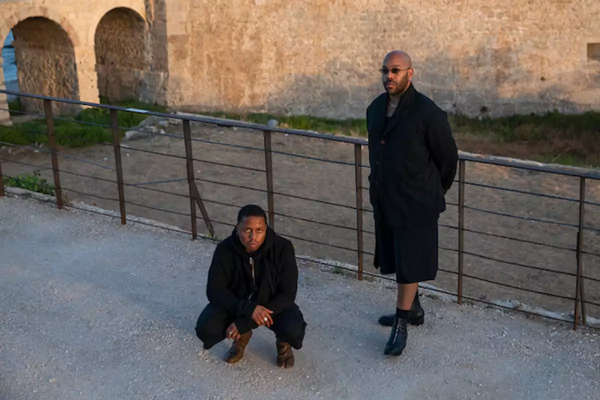 Two people dressed in all black – one stood up, one squatting next to metal railing at sunset