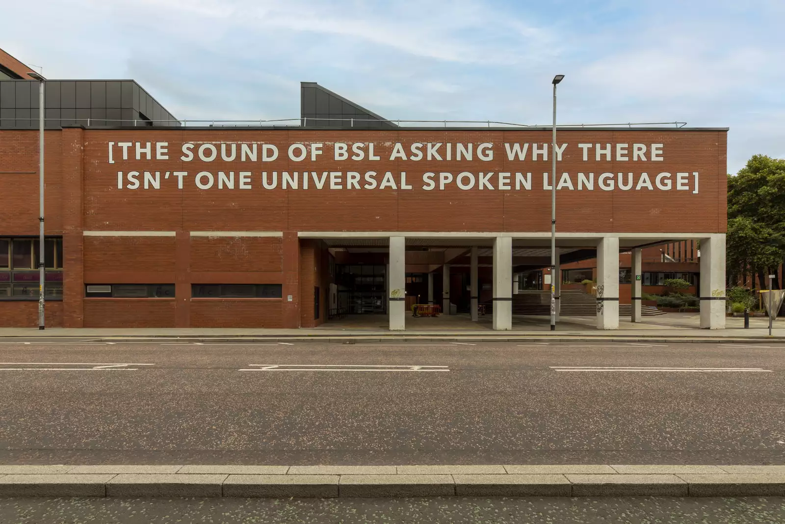 A photograph of a building with a large text installation applied with white adhesive vinyl. The text reads "[THE SOUND OF BSL ASKING WHY THERE ISN'T ONE UNIVERSAL SPOKEN LANGUAGE".