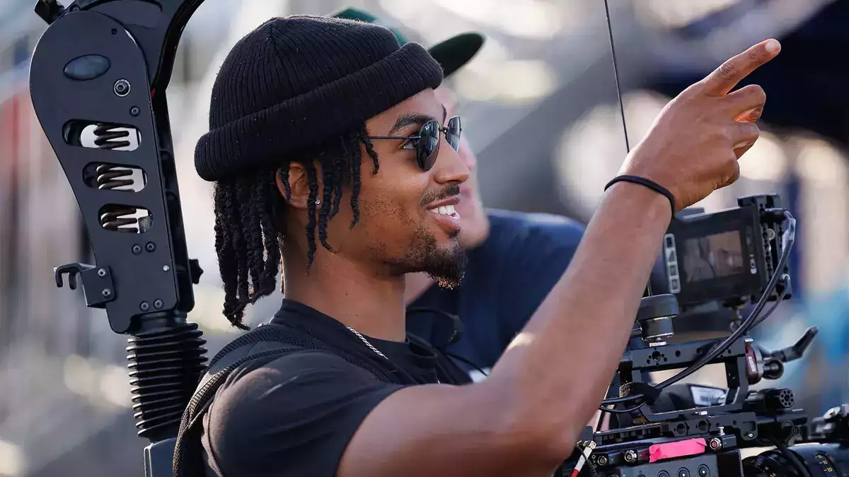 A young Black man wearing sunglasses and a beanie hat operates a video camera.