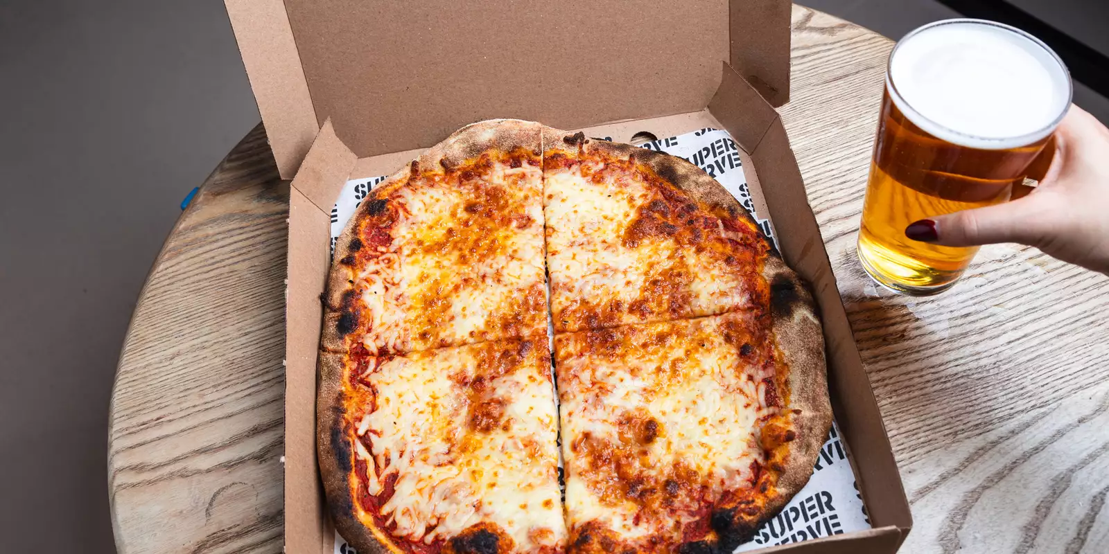 An open pizza box on a wooden table with a cheese pizza cut into four slices. A hand with red-painted nails holds a pint of beer beside it.