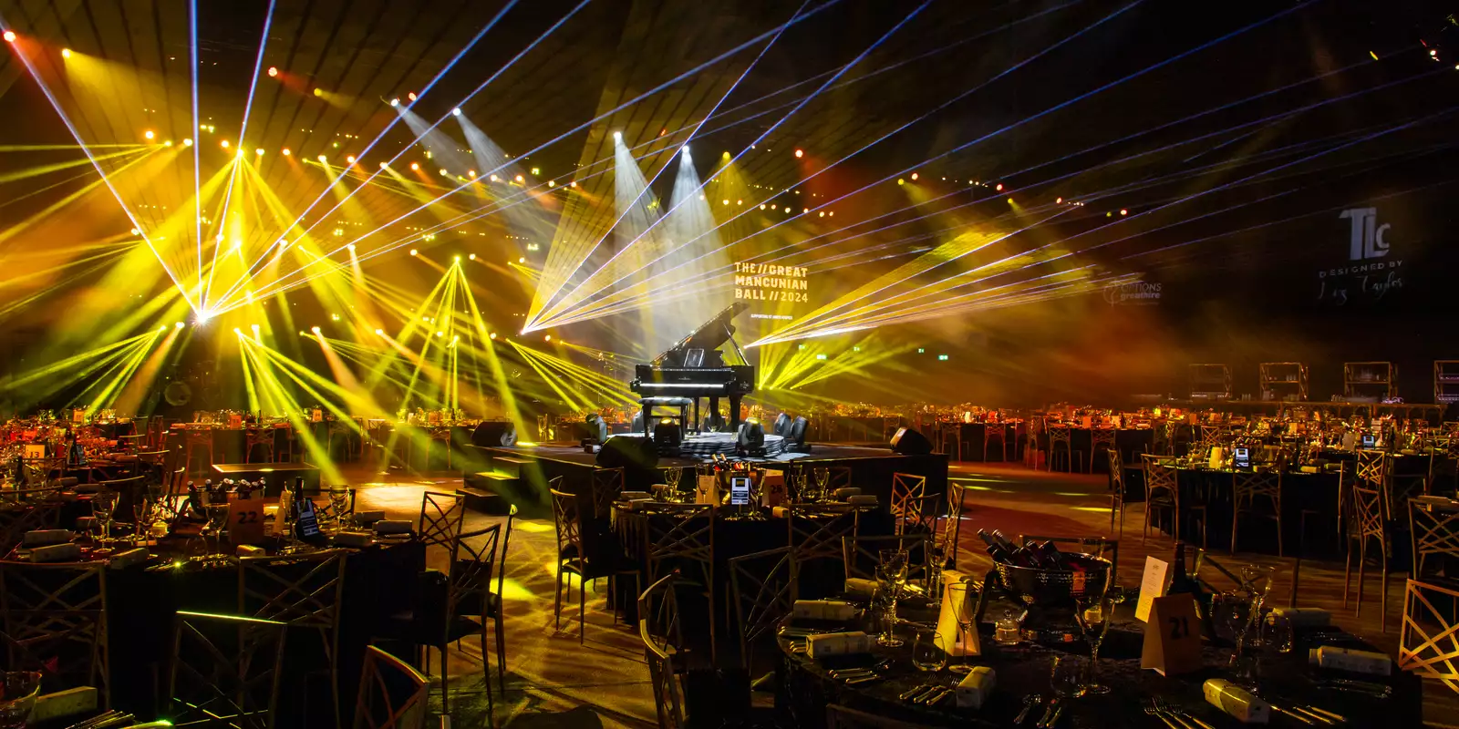 Large circle tables set up in the Warehouse for the Great Mancunian Ball. There are yellow laser lights and a grand piano in the centre.