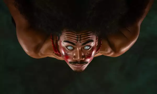 A member of FAFSWAG with glowing white eyes and red and black face paint stares intensely at the camera. They wear red beaded necklaces, and the image is flipped upside down.