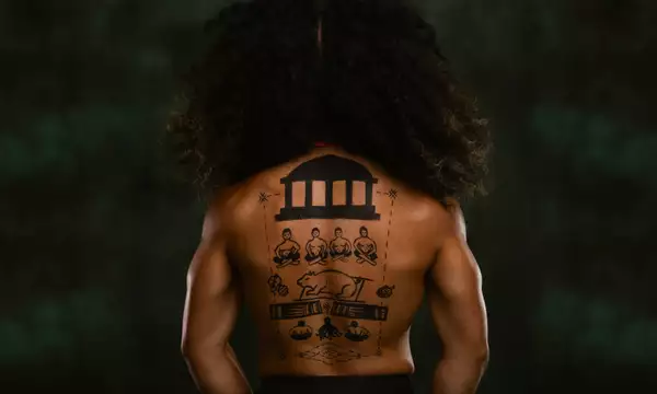 A member of FAFSWAG with their back turned away from the camera. They are topless with hand-drawings on their back. The background is dark green.