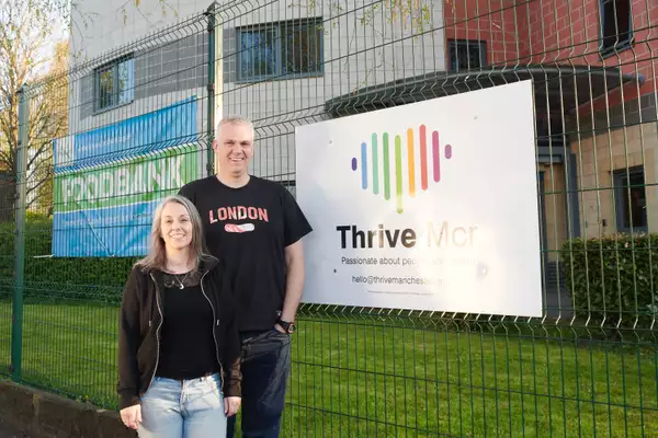 Two people stood outside in front of a metal fence, wearing black tops and jeans. They are stood next to a sign which says 'Thrive MCR: passionate about people and health'