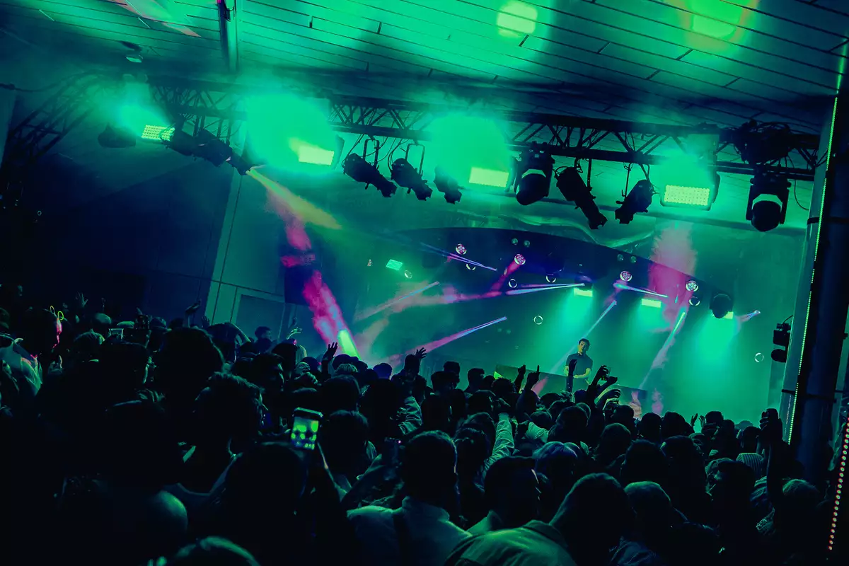 A DJ set on Festival Square from MIF23, showing a large crowd of people dancing and filming a DJ on stage. The lighting is hazy green and there are purple strobe lights.