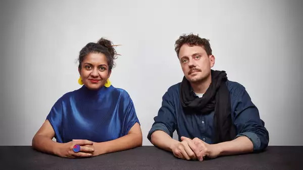 Two members of Superflux sat at a table, looking at the camera. The person on the left is wearing a shiny blue top and yellow earrings. The person on the right is wearing a blue shirt and black scarf.