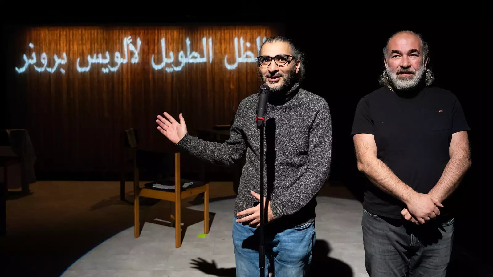 On stage, two people speak into a microphone with Arabic writing on the wall behind them