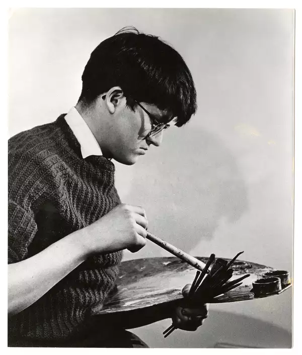 Black and white photograph of a young David Hockney holding an easel. He is wearing a jumper, shirt and glasses.