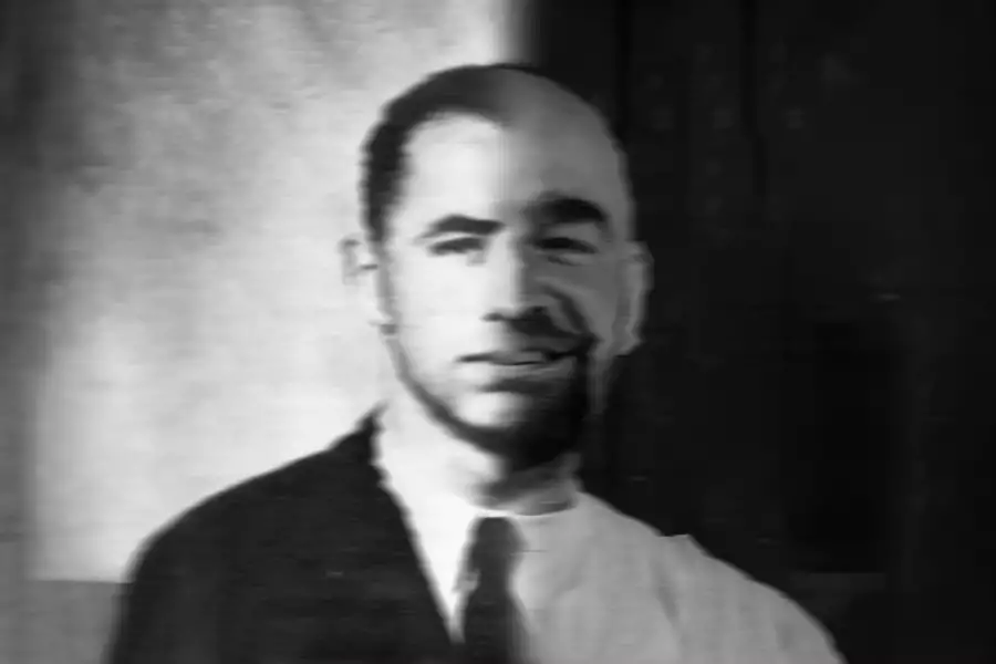 Black and white photo headshot of a person. One half of their body is clean-shaven and wearing a suit and tie; the other half is not.