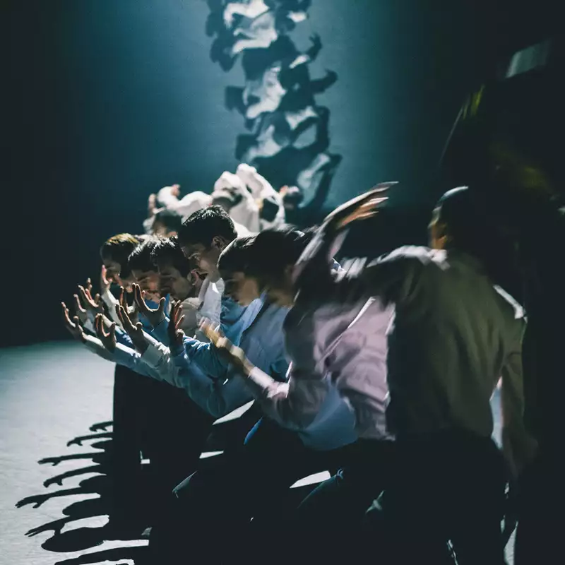 A group of dancers stood in a line with their arms extended forward, moving in blurred motion. The lighting is dim, casting dramatic shadows.