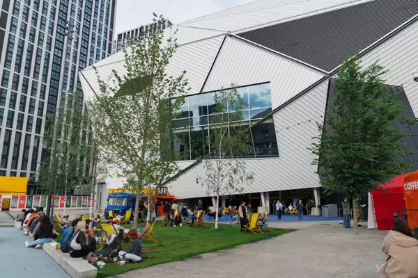 Groups of people sat outside Aviva Studios on the grass during MUBI FEST. There are yellow deck chairs plus a bar truck and coffee cart.