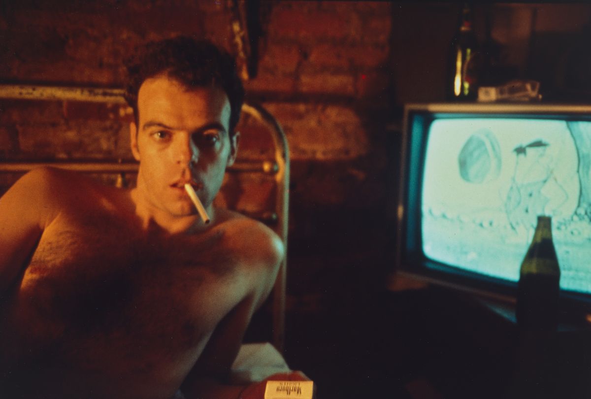Friend of artist Nan Goldin laying on a bed with a cigarette in his mouth while the Flintstones are running on a television.