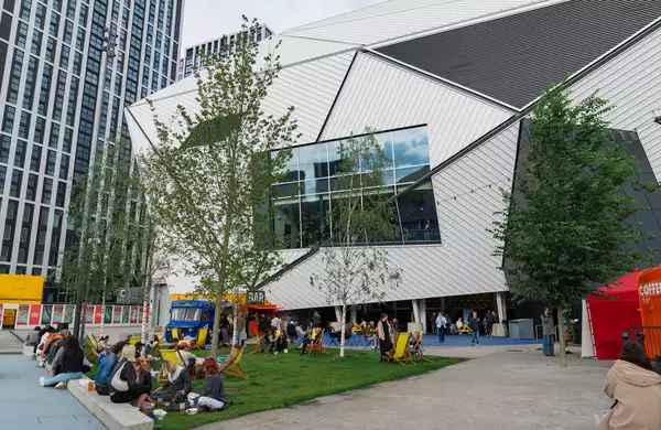 Groups of people sat outside Aviva Studios on the grass during MUBI FEST. There are yellow deck chairs plus a bar truck and coffee cart.