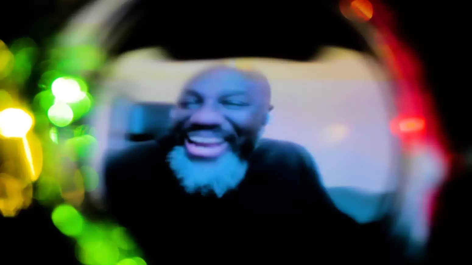 A blurred and light-distorted image down a dark camera lens of a laughing person who has black skin, a bald head and a bushy black and white beard.