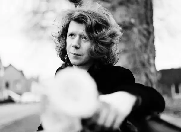 A black and white photograph of Factory Sounds 2023 cohort member James Holt sat on a bench in front of a tree trunk