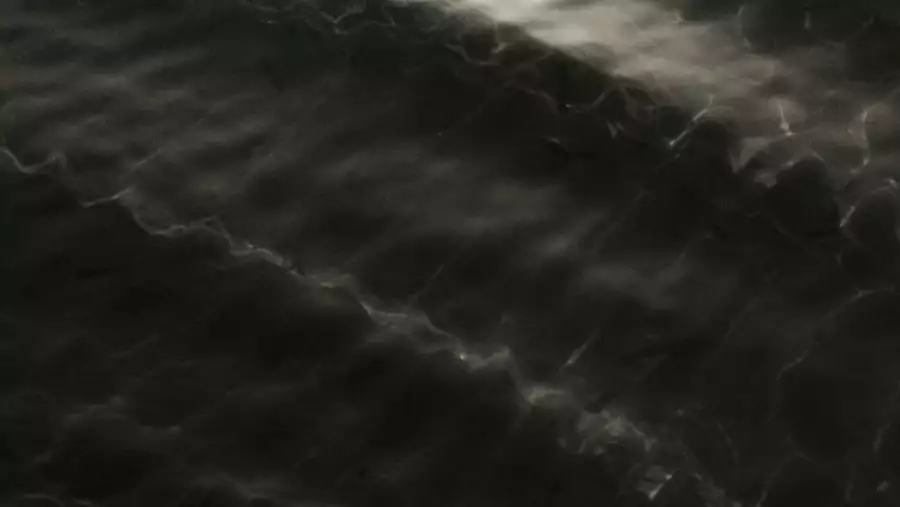 This image shows a dark, wavy surface with subtle reflections, resembling the texture of water ripples or light passing through water.
