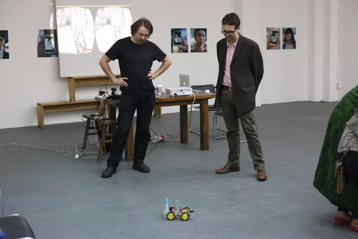 Josh Harle and another person observing a wheeled robot on the ground.