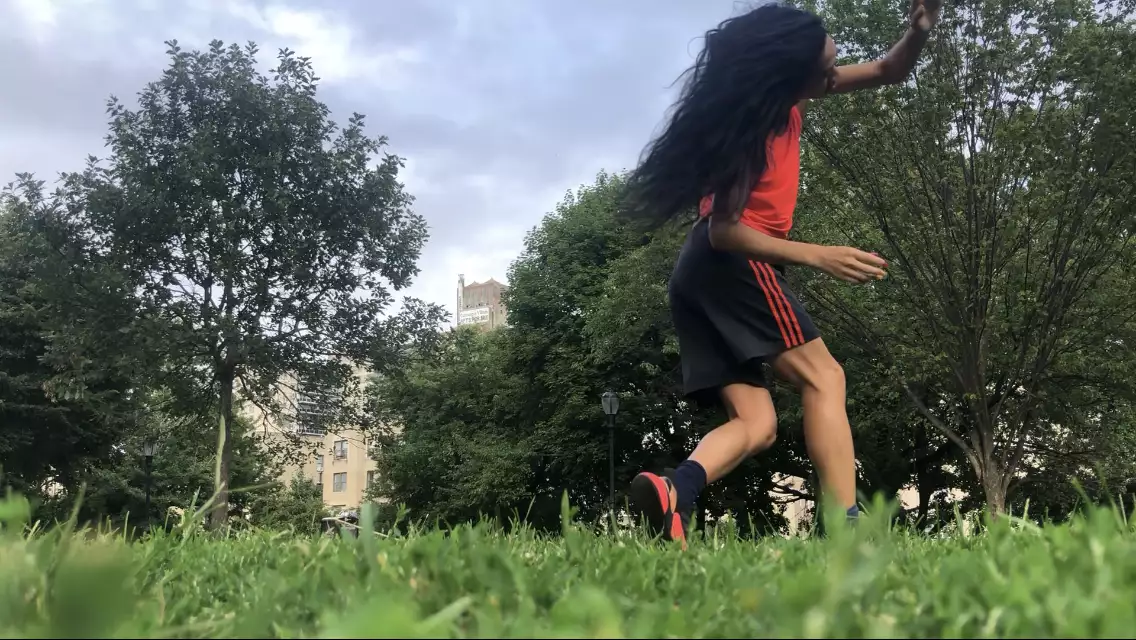 A horizontal photo of ​​Benedict Nguyễn, wearing black shorts, orange tank, and black and orange sneakers, lunging forward on right leg with left arm reaching across her face, in a field with green grass and trees, with buildings in distant background.