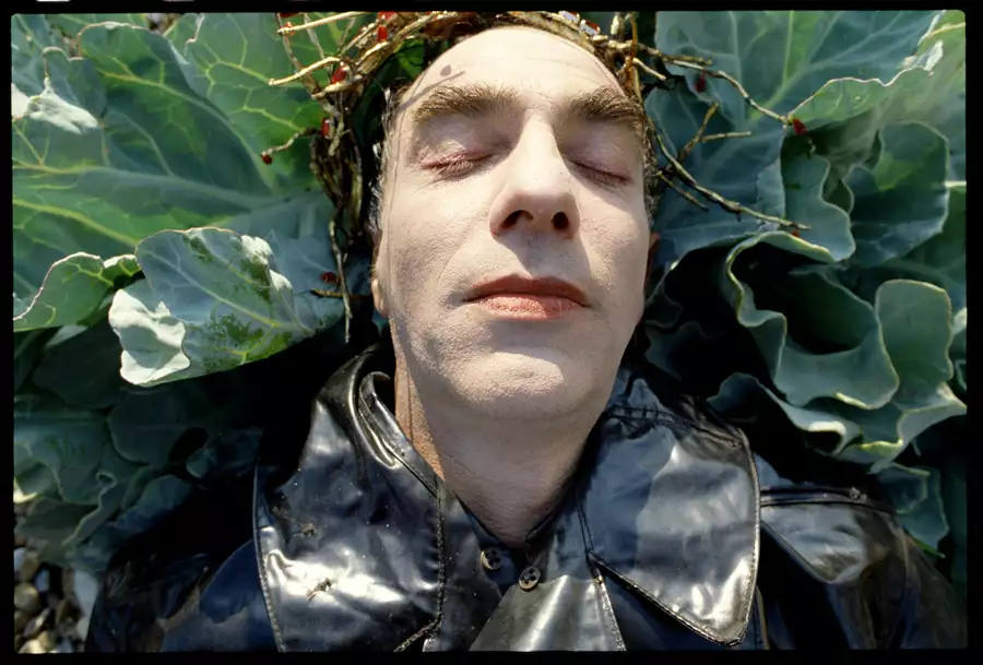 Still from Derek Jarman's The Garden (1990), showing a close up of Jarman's face. He is lying on a cabbage plant, wearing a black leather jacket and golden crown.