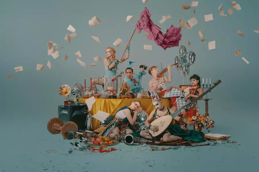 A group of performers around a table littered with objects, some are playing musical instruments, one is waving a flag
