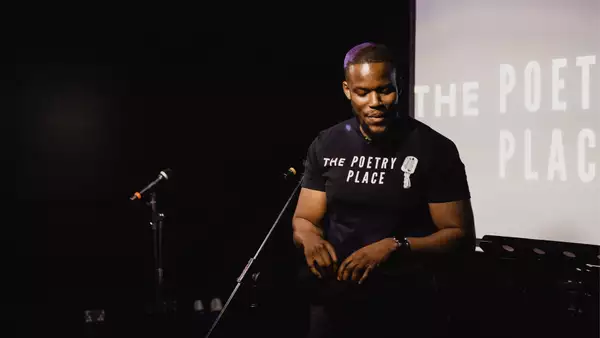 Man on stage performing, with a tee shirt that read The Poetry Place