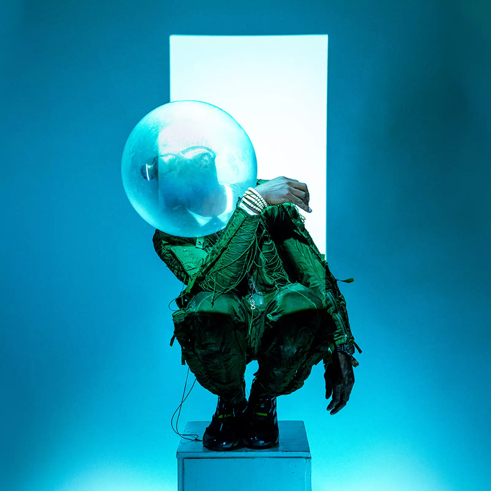 Benji Reid crouches on a pedestal, wearing a textured green outfit and a large transparent bubble helmet. There is blue lighting, with a glowing white rectangle in the background.
