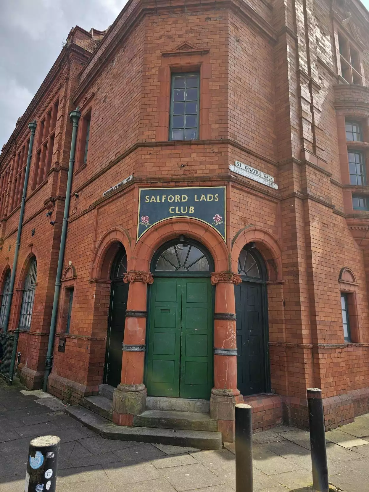 The historic red-brick facade of Salford Lads Club, with green doors and ornate archways, under a cloudy sky.