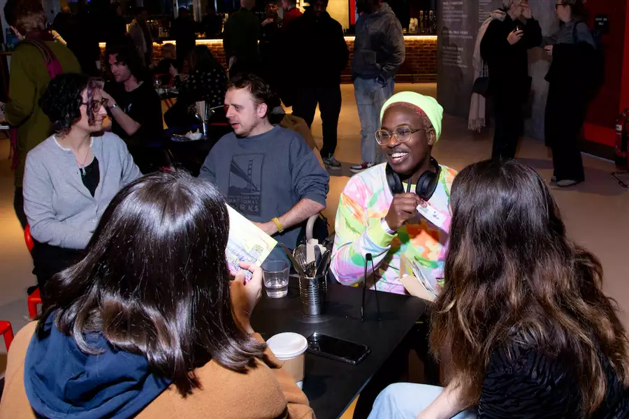 A group of people sat around a table in the Social, engaged in conversation and laughter. The table is scattered with drinks, papers, and personal items. In the background are other people chatting.