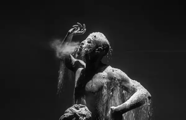 A dramatic black-and-white photograph of a man covered in dust and sand, with particles cascading over his body as he exhales forcefully.